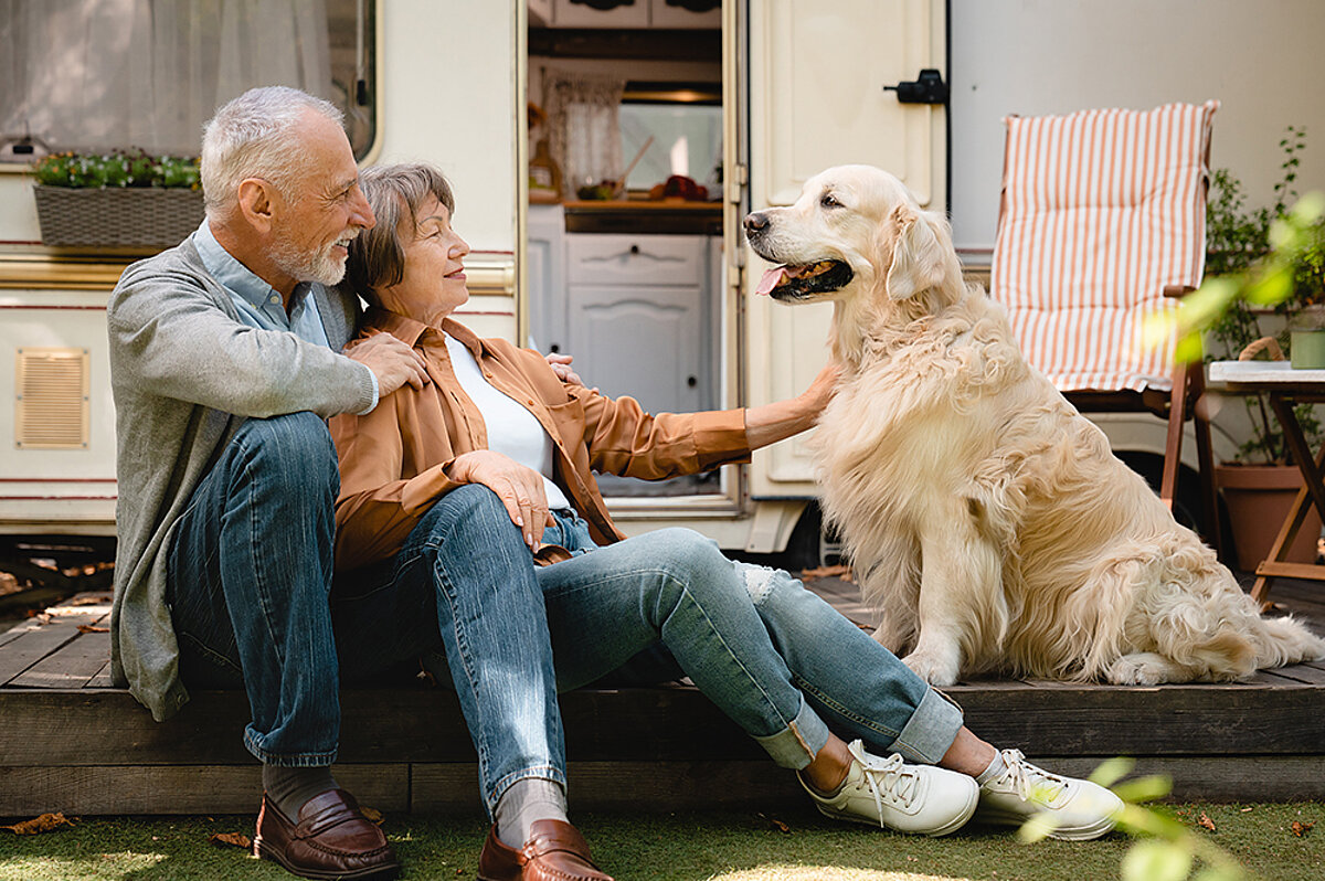 Zwei Personen sitzen mit einem großen, hellen Hund vor einem Wohnmobil.