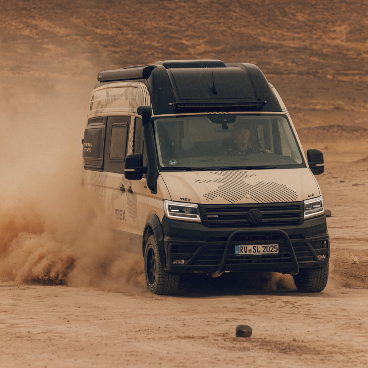 Ein heller Camper Van von vorne fährt durch ein wüstenähnliches Gelände. 