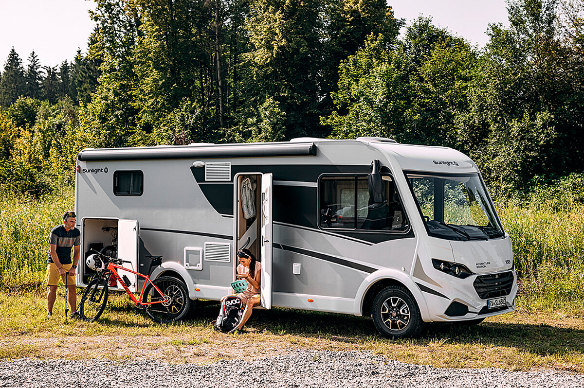 Ein helles Wohnmobil steht auf einem Platz mit Wald im Hintergrund und Menschen davor.